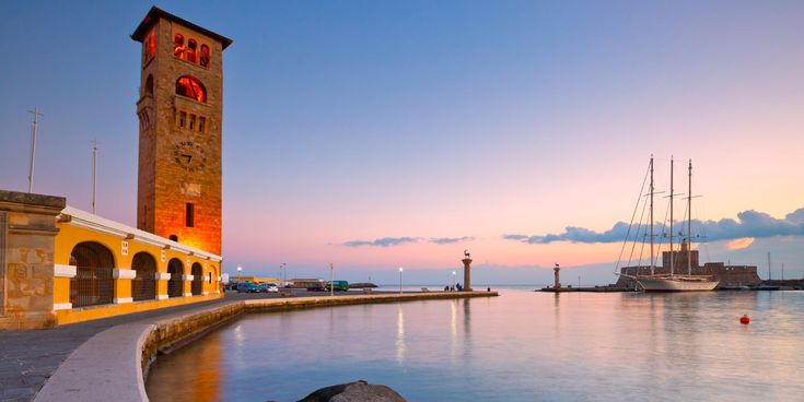 Hafen von Rhodos-Stadt bei Sonnenuntergang mit Glockenturm, Segelschiff und den Säulen mit Hirschstatuen am Wasser.