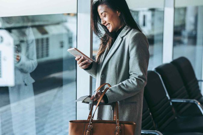 Geschäftsreisende lächelt auf ihr Smartphone im Flughafen-Wartebereich.