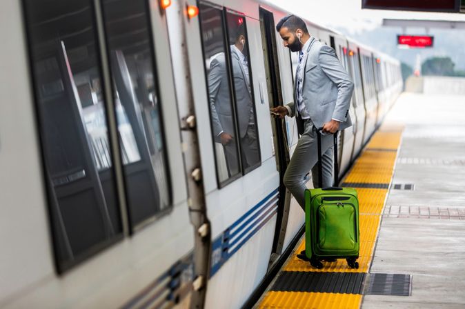 Geschäftsmann im grauen Anzug mit grünem Rollkoffer steigt in einen Zug, dabei schaut er auf sein Smartphone.