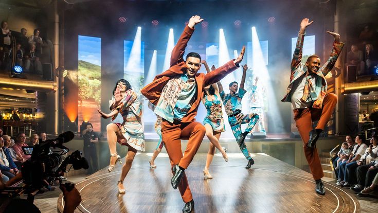 Eine Gruppe von Tänzerinnen und Tänzern in bunten Kostümen führt auf einer Bühne vor Publikum eine energiegeladene Show mit ausdrucksstarken Sprüngen und Bewegungen auf.