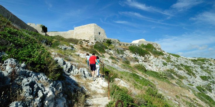 Wandernde steigen über einen felsigen Pfad zu einer historischen Festungsanlage auf einem Hügel hinauf.
