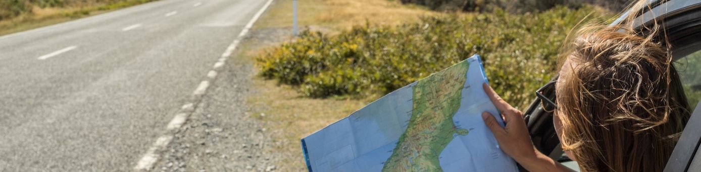 Reisende schaut aus dem Auto und hält eine Landkarte in der Hand, während eine Straße durch eine Berglandschaft führt – Sinnbild für individuelle Erkundungen mit dem Mietwagen.