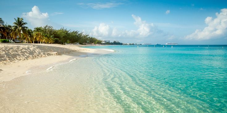 Weißer Sandstrand mit kristallklarem Wasser, im Hintergrund sind man vage ein paar Kreuzfahrtschiffe am Hafenanleger