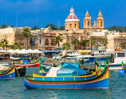 Traditionelle bunte Luzzus schwimmen im Hafen von Marsaxlokk auf Malta, vor einer Kulisse aus Palmen, Cafés und einer Kirche mit roter Kuppel und Doppelturm.