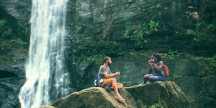 Zwei Reisende sitzen auf Felsen vor einem Wasserfall, machen eine Pause und fotografieren die beeindruckende Naturkulisse.