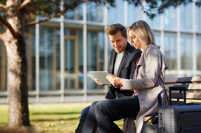 Geschäftsreisende (Mann und Frau) auf einer Bank vor einem Bürogebäude mit einem Tablet in der Hand