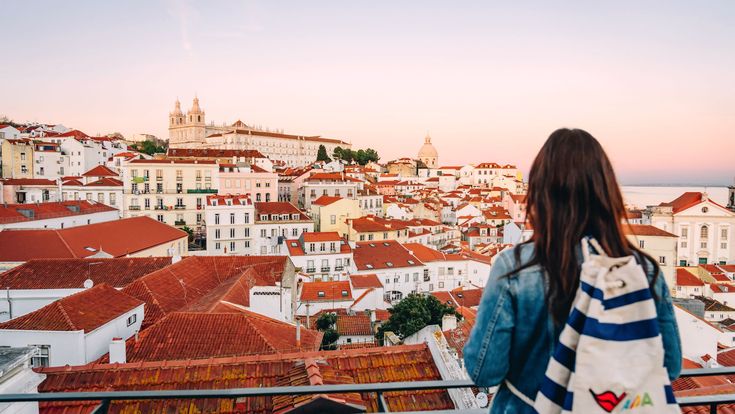 Eine Frau mit Rucksack blickt bei Sonnenuntergang von einer Aussichtsterrasse auf die Altstadt mit roten Dächern und historischen Gebäuden.