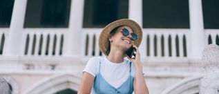 Lächelnde Urlauberin mit Strohhut und Sonnenbrille telefoniert vor einem weißen Gebäude mit Säulen.