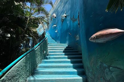 Blaue Treppe mit türkisfarbenem Geländer, rechts Wand mit plastischen Fischen und Meeresmotiven, links Palmen und blauer Himmel