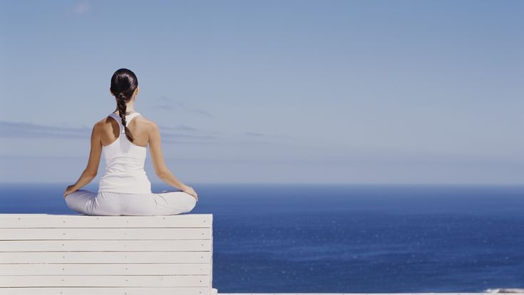 Eine Frau sitzt im Schneidersitz auf einer weißen Terrasse und meditiert mit Blick auf das ruhige, blaue Meer unter klarem Himmel.
