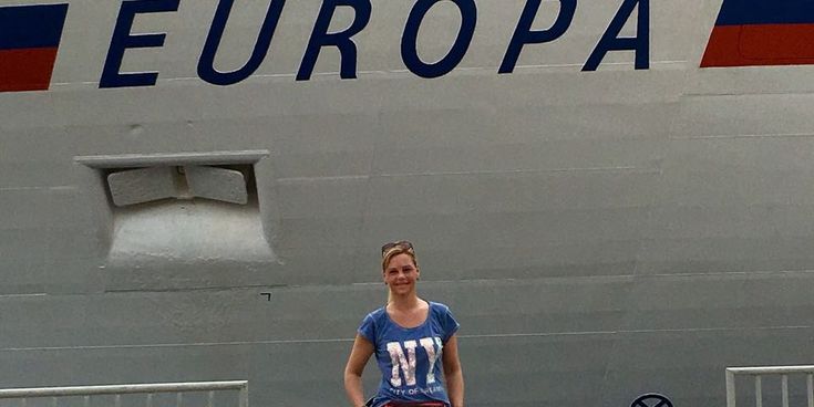 Christina steht vor dem Kreuzfahrtschiff MS Europa am Hafen, der Schiffsname ist gut sichtbar am Rumpf.