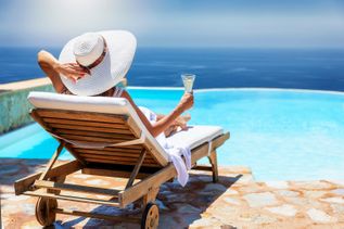 Frau mit Hut und Cocktail auf Sonnenlieg am Pool mit Blick über das Meer