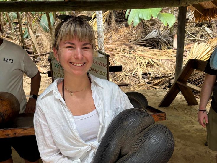 Georgina Dörling hält lächelnd eine große Coco-de-Mer-Nuss in einem tropischen Garten auf den Seychellen.
