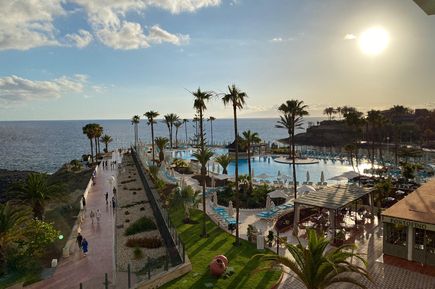 Blick auf Hotelpool mit Liegestühlen und Palmen, Promenade am Meer bei Sonnenuntergang unter bewölktem Himmel.