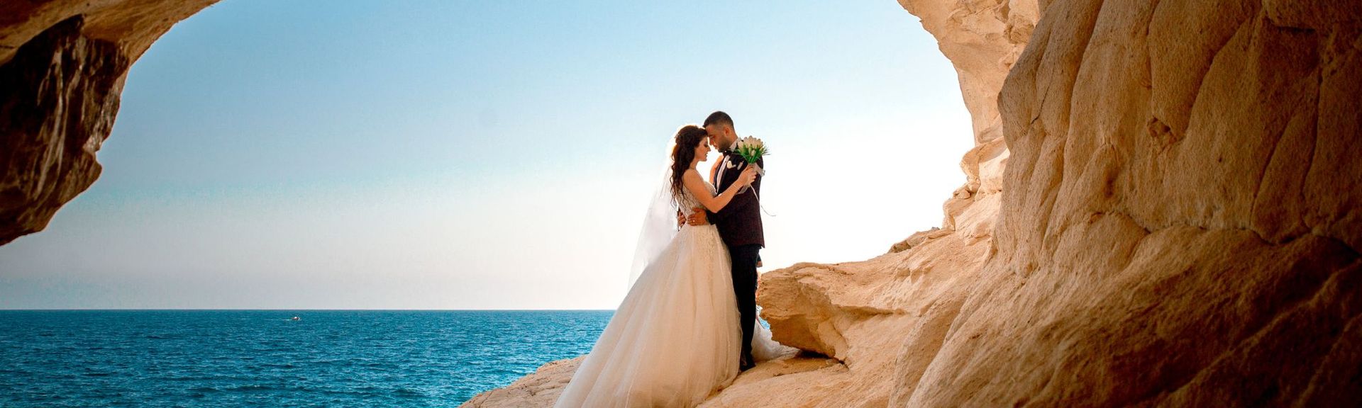 Brautpaar steht in einer Felsgrotte mit Blick auf das Meer, umgeben von hellem Sandstein und blauem Himmel – Symbol für Liebe und Verbundenheit.