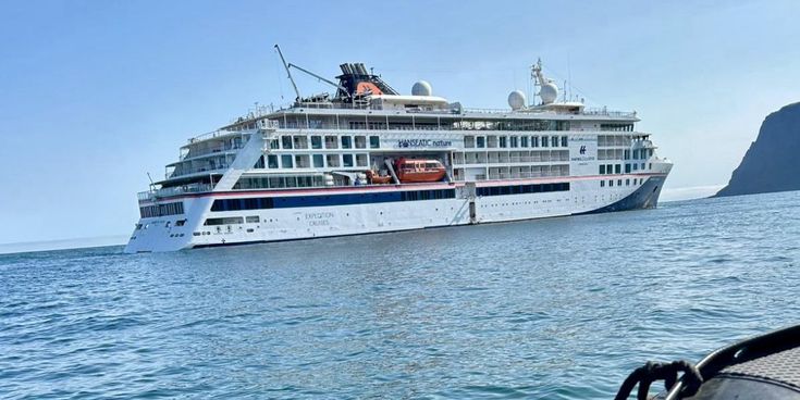 Blick vom Schlauchboot auf ein modernes Expeditionskreuzfahrtschiff, das vor der Küste auf ruhigem, türkisfarbenem Meer ankert, bei klarem Himmel und felsiger Küstenlandschaft im Hintergrund.