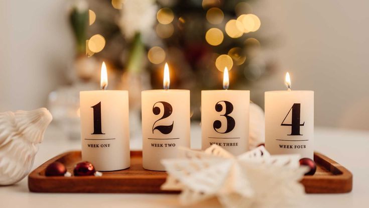 Vier brennende Adventskerzen mit den Zahlen eins bis vier stehen auf einem Holztablett vor einem festlich beleuchteten Weihnachtsbaum und symbolisieren die besinnliche Vorweihnachtszeit.