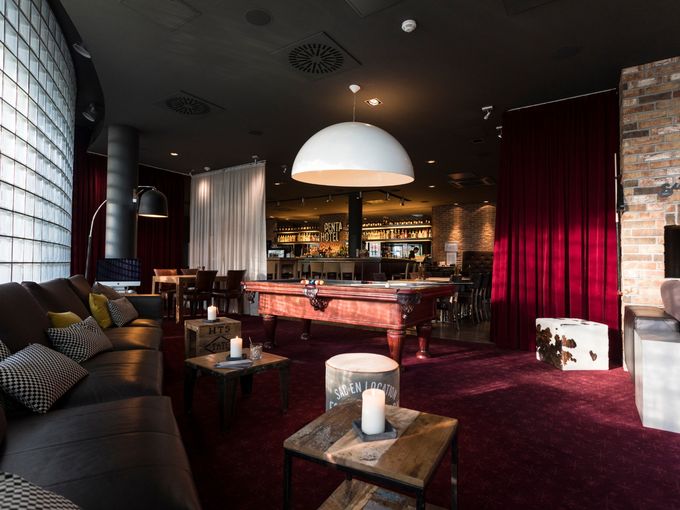 Gemütliche Hotellounge des Penta Hotels mit Ledersofas, Billardtisch, Bar im Hintergrund und stimmungsvoller Beleuchtung in warmen Rottönen