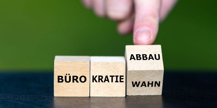 Eine Hand dreht den dritten von drei nebeneinanderstehenden Holzwürfeln um. Die Würfel bilden zusammen entweder das Wort „Bürokratiewahn" oder „Bürokratieabbau" – je nach Stellung des dritten Würfels. Hintergrund ist grün und unscharf.