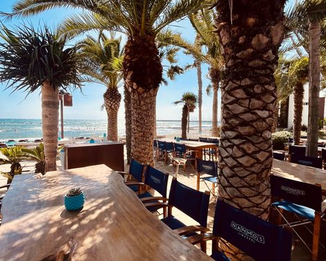 Sonnige Strandterrasse mit Holztisch, blauer Dekoration, Stühlen mit blauer Rückenlehne und Palmen im Hintergrund.