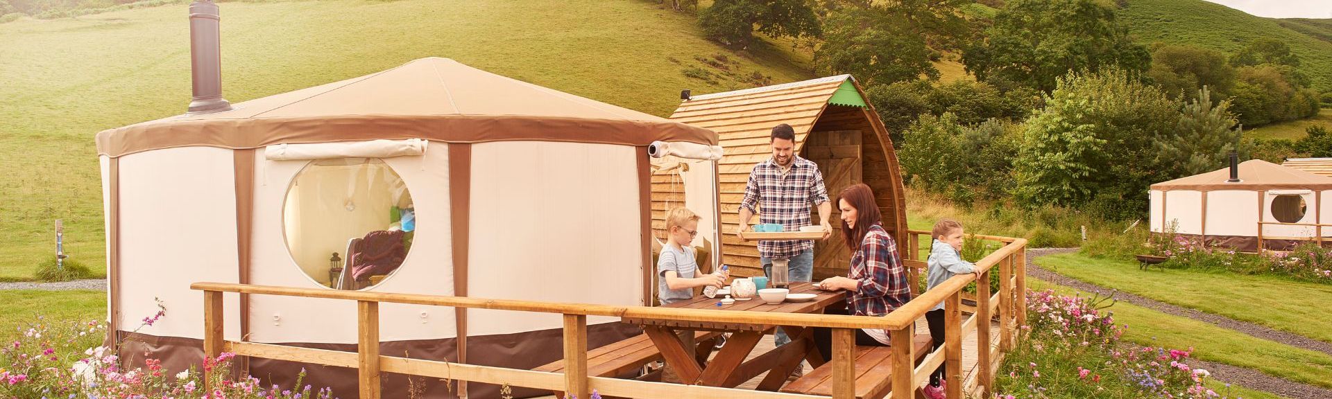 Eine Familie genießt ein Mittagessen auf der Holzterrasse vor einem Glamping-Zelt inmitten grüner Hügel und Blumenbeeten.