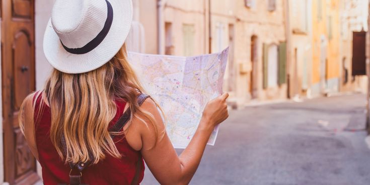Reisende Frau mit Hut und Rucksack steht in einer sonnigen Altstadtgasse und schaut auf eine Stadtkarte.