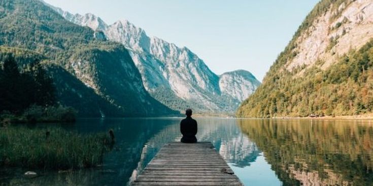 Person sitzt ruhig auf einem Holzsteg, der in einen klaren Bergsee führt, umgeben von imposanten Gipfeln – Moment der Stille und Verbundenheit mit der Natur.