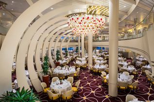 Ein eleganter Speisesaal auf einem Kreuzfahrtschiff mit geschwungenen Deckenelementen, festlich gedeckten Tischen und einem großen, kunstvollen Kronleuchter.