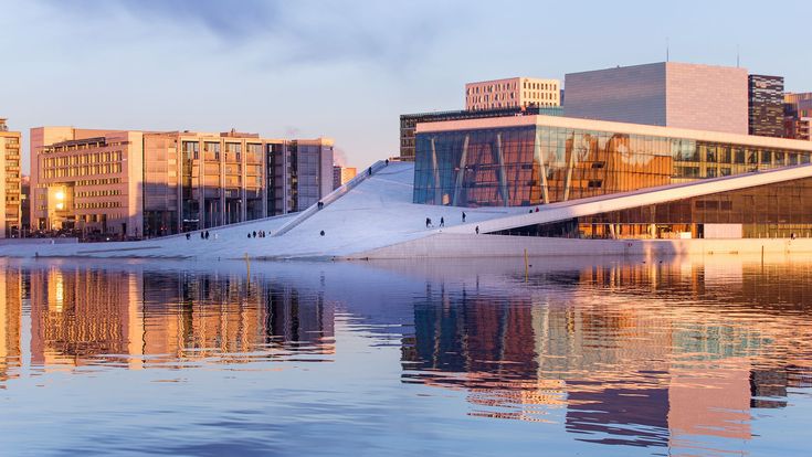 Das moderne Osloer Opernhaus mit schräger begehbarer Dachfläche spiegelt sich im ruhigen Wasser, während Menschen bei Sonnenuntergang darauf spazieren.