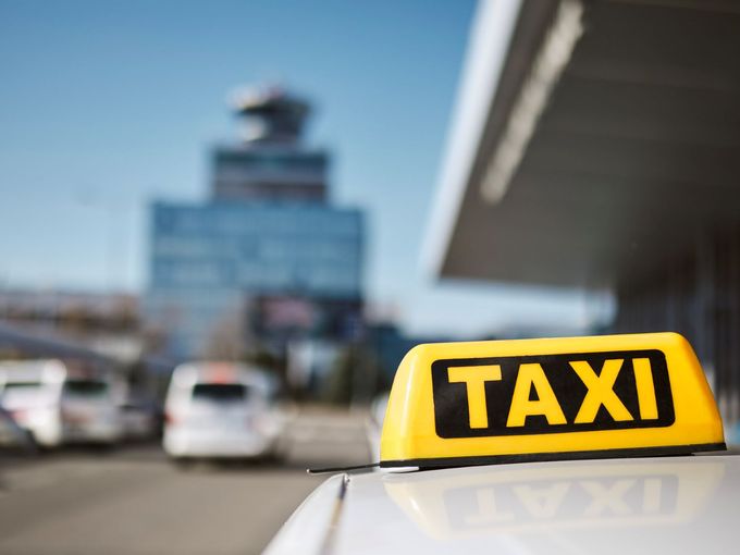 Nahaufnahme eines gelb-schwarzen Taxischilds auf dem Dach eines weißen Taxis. Im unscharfen Hintergrund sind ein Flughafengebäude mit Kontrollturm sowie weitere Fahrzeuge auf einem Parkplatz zu sehen.