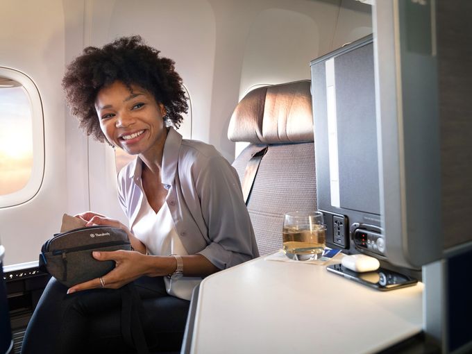 Lächelnde Frau sitzt komfortabel in der Business Class eines Flugzeugs mit einem Getränk und einer Reisetasche.