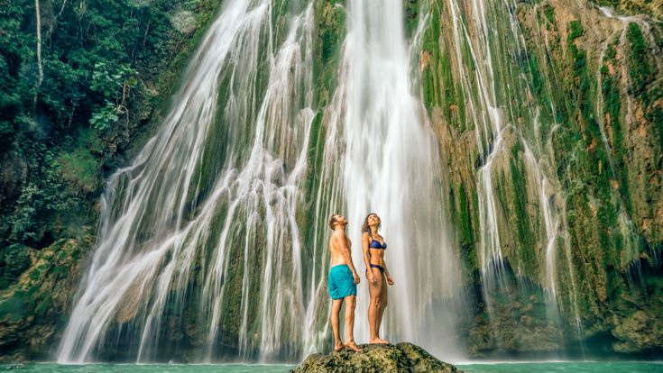 Ein Paar in Badekleidung steht auf einem Felsen vor einem hohen Wasserfall, der über grün bewachsene Felsen in ein türkisfarbenes Becken stürzt.