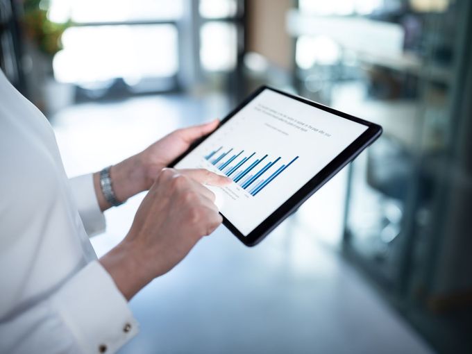 Eine Person in weißem Hemd hält ein Tablet in beiden Händen und tippt mit dem Zeigefinger auf ein Balkendiagramm, das auf dem Display zu sehen ist. Im Hintergrund ein heller, verschwommener Büroflur.