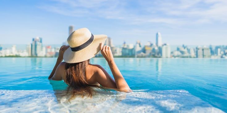 Frau mit Strohhut steht in einem Infinitypool und blickt auf eine moderne Großstadtskyline.