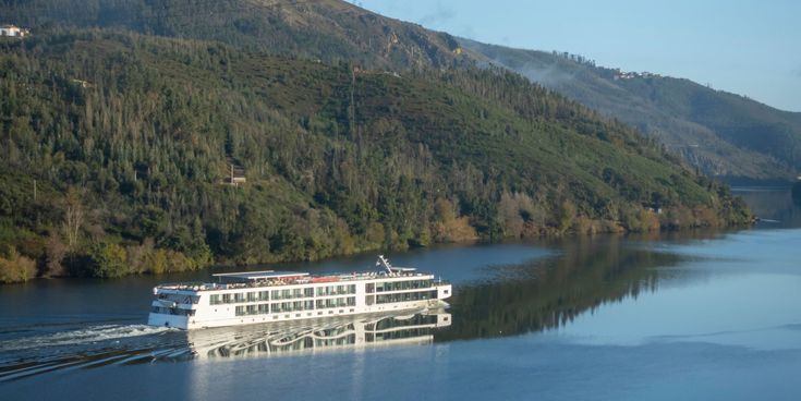 Weißes Flusskreuzfahrtschiff fährt durch das ruhige Wasser des Douro-Flusses, umgeben von grünen Hügeln und bewaldeten Berghängen.