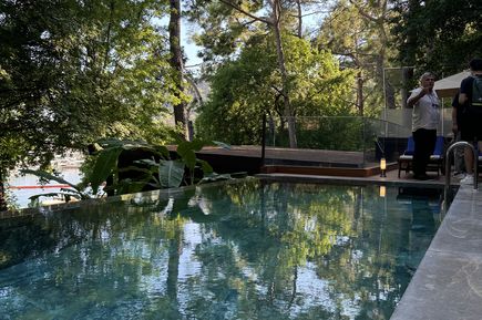 Schwimmbecken mit klarem Wasser, umgeben von Bäumen und einer Terrasse mit zwei Personen am Beckenrand.