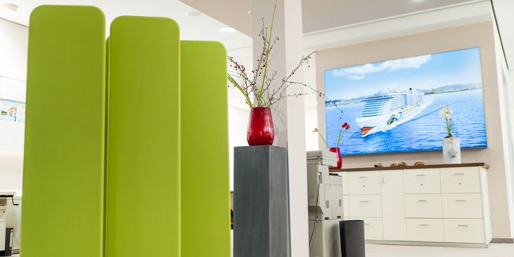 Innenraum des Reisebüros mit grüner Raumtrennung, roter Blumenvase und einem großen Bildschirm mit Kreuzfahrtmotiv im Hintergrund.