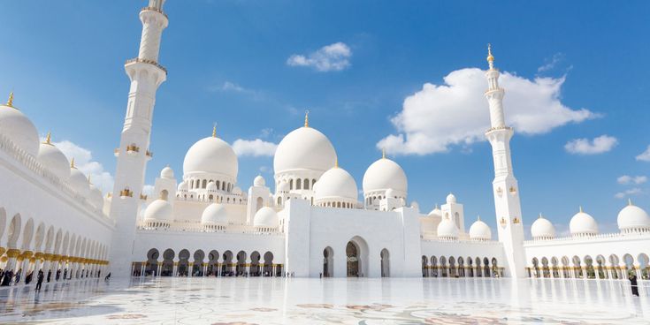 Weiße Kuppeln und Minarette der Sheikh-Zayed-Moschee in Abu Dhabi vor blauem Himmel mit verziertem Marmorboden im Vordergrund.