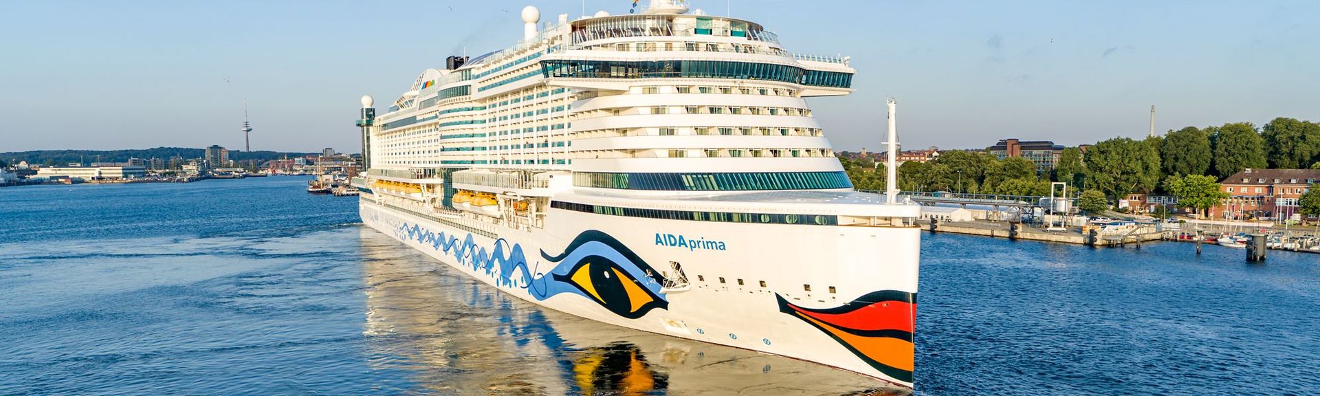 Das Kreuzfahrtschiff AIDAprima fährt bei sonnigem Wetter durch einen Hafen mit Stadt- und Uferkulisse im Hintergrund.