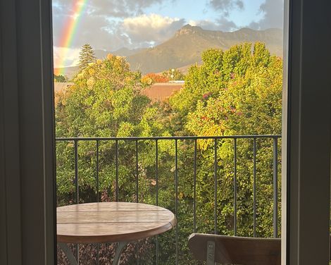 Blick vom Balkon auf einen leuchtenden Regenbogen über grünen Gärten und den Bergen bei Stellenbosch.