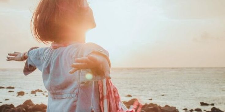 Frau steht mit ausgebreiteten Armen am Meer und genießt den Sonnenuntergang – Symbol für Freiheit, Leichtigkeit und das Gefühl von Urlaub.