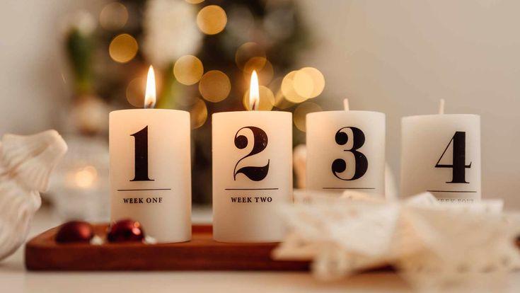 Vier brennende Adventskerzen mit den Zahlen eins bis vier stehen auf einem Holztablett vor einem festlich beleuchteten Weihnachtsbaum und symbolisieren die besinnliche Vorweihnachtszeit.