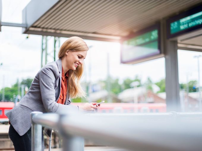 Geschäftsfrau steht lächelnd am Bahnhof und schaut auf ihr Smartphone