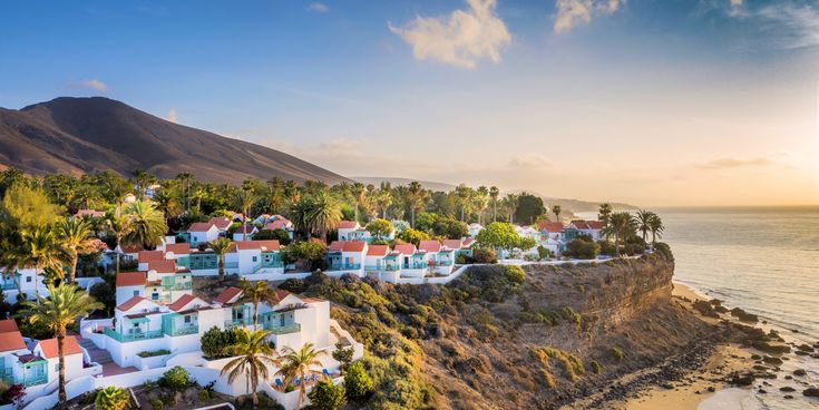 Küstenort auf Fuerteventura mit weißen Häusern und roten Dächern auf einer Klippe über dem Meer, umgeben von Palmen und Bergen im Hintergrund.