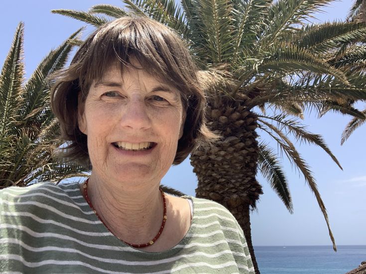 Petra Meyers lächelt vor einer großen Palme und dem blauen Meer auf Fuerteventura.